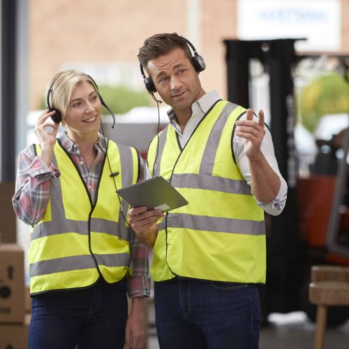 Male And Female Workers Wearing Headsets In Logistics Distribution Warehouse Using Digital Tablet